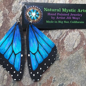 May include: A pair of handmade butterfly wing earrings. The wings are painted with shades of blue and black, with white dots along the edges. The earrings are made by Natural Mystic Arts, hand painted by Artist Jill Woyt, and made in Big Sur, California.