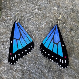 May include: A pair of butterfly wing earrings. Each earring features a blue and turquoise wing design with black edges and white dots. The earrings have silver-colored hooks.