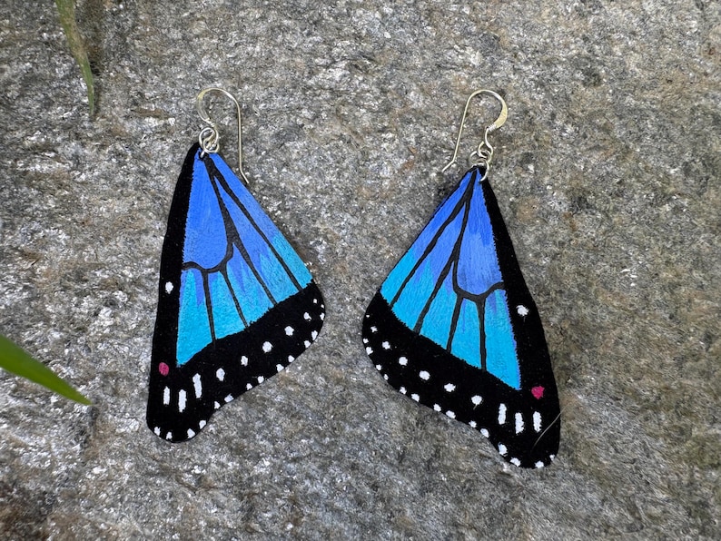 May include: A pair of butterfly wing earrings. Each earring features a blue and turquoise wing design with black edges and white dots. The earrings have silver-colored hooks.