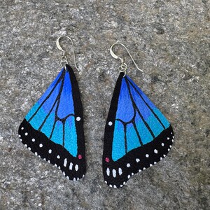 May include: A pair of butterfly wing earrings. Each earring features a blue and turquoise wing design with black edges and white dots. The earrings have silver-colored hooks.
