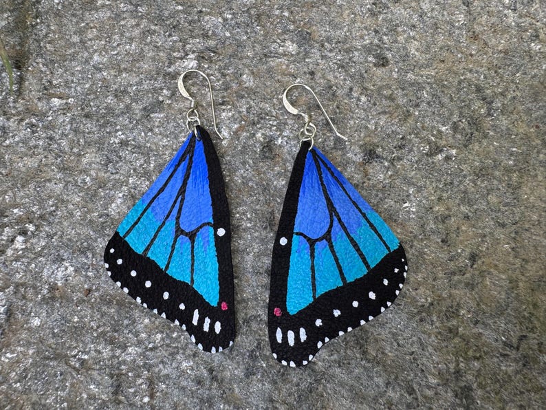 May include: A pair of butterfly wing earrings. Each earring features a blue and turquoise wing design with black edges and white dots. The earrings have silver-colored hooks.