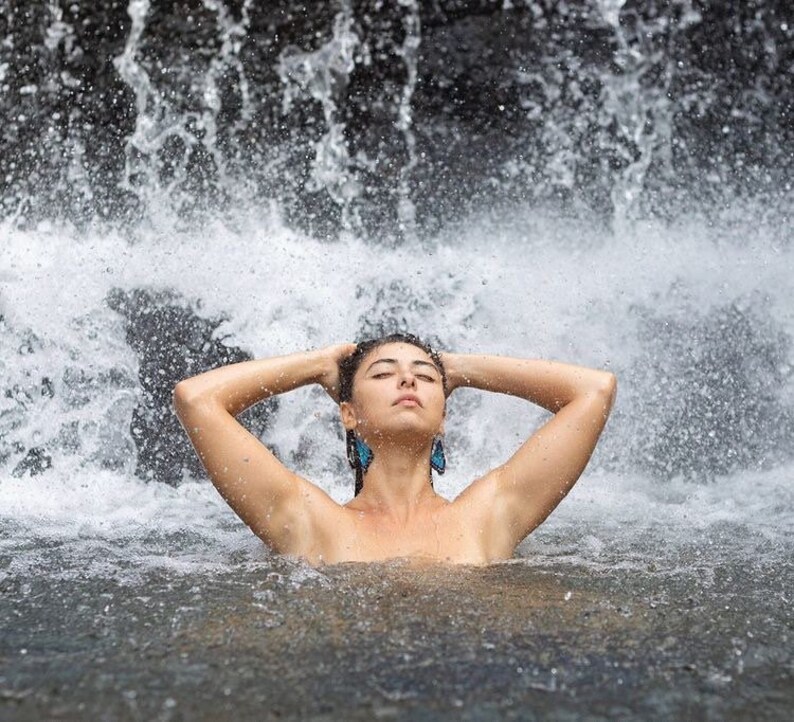 May include: A person stands in a waterfall, with arms raised to the head. The person wears blue butterfly earrings. Water cascades down in the background, creating a refreshing and natural scene.