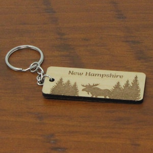 May include: Wooden keychain with a moose silhouette and the text "New Hampshire" engraved on it. The keychain is brown and has a silver key ring.
