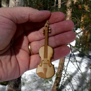 May include: A wooden violin ornament with a natural wood finish. The ornament is hanging from a twine string.