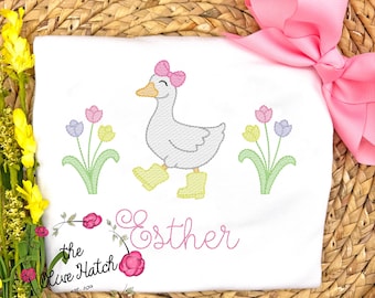 May include: White embroidered baby onesie with a pink bow, a gray duck wearing yellow rain boots, and three pink, yellow, and purple flowers. The onesie has the name "Esther" embroidered in pink.