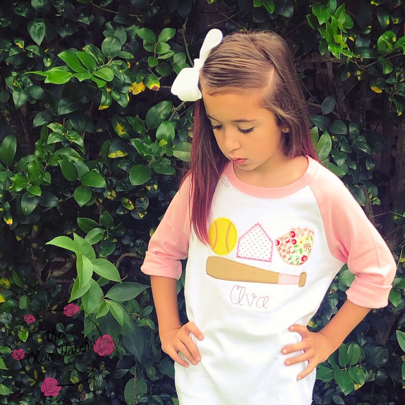 Puede incluir: Una camiseta de b&eacute;isbol de color blanco y rosa con un dise&ntilde;o de pelota de softball, home plate y bate de b&eacute;isbol. La camiseta tiene el nombre "Ava" bordado en la parte delantera.