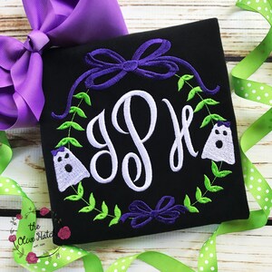 Peut inclure: T-shirt noir carr&eacute; avec un monogramme blanc, entour&eacute; d'une couronne de feuilles vertes, de n&oelig;uds violets et de deux fant&ocirc;mes brod&eacute;s. Un grand n&oelig;ud violet et un ruban vert &agrave; pois sont en arri&egrave;re-plan. Le logo Olive Hatch est en bas &agrave; gauche.