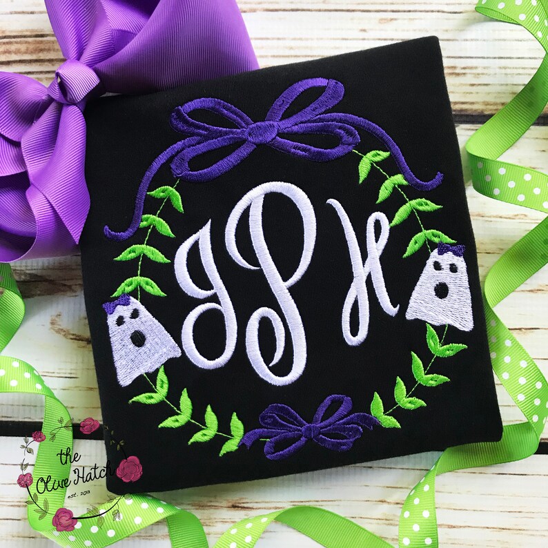 Peut inclure: T-shirt noir carr&eacute; avec un monogramme blanc, entour&eacute; d'une couronne de feuilles vertes, de n&oelig;uds violets et de deux fant&ocirc;mes brod&eacute;s. Un grand n&oelig;ud violet et un ruban vert &agrave; pois sont en arri&egrave;re-plan. Le logo Olive Hatch est en bas &agrave; gauche.