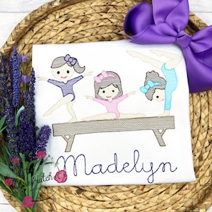 Puede incluir: Dise&ntilde;o bordado de tres ni&ntilde;as haciendo gimnasia en una viga de equilibrio. Las ni&ntilde;as llevan leotardos y tienen lazos en el pelo. El dise&ntilde;o est&aacute; en una camiseta blanca con el nombre "Madelyn" bordado debajo de la viga.