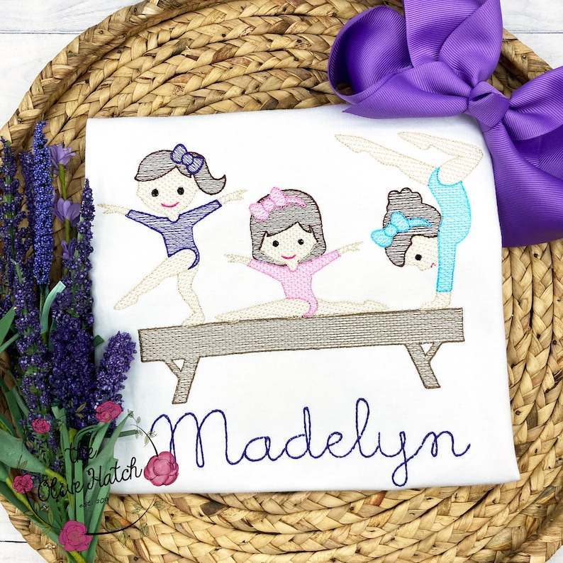 Puede incluir: Dise&ntilde;o bordado de tres ni&ntilde;as haciendo gimnasia en una viga de equilibrio. Las ni&ntilde;as llevan leotardos y tienen lazos en el pelo. El dise&ntilde;o est&aacute; en una camiseta blanca con el nombre "Madelyn" bordado debajo de la viga.