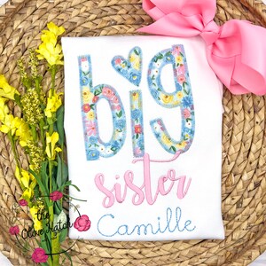 May include: White t-shirt with the words "Big Sister" in pink and blue floral letters. The name "Camille" is embroidered in blue below the text.