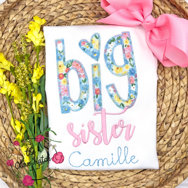 May include: White t-shirt with the words "Big Sister" in pink and blue floral letters. The name "Camille" is embroidered in blue below the text.