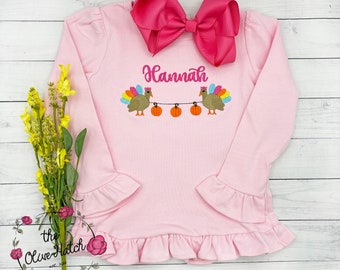 May include: A light pink long-sleeved shirt with ruffled cuffs and hem. The shirt features an embroidered design of two turkeys, pumpkins, and the name "Hannah" in pink. A large pink bow is attached at the neckline.