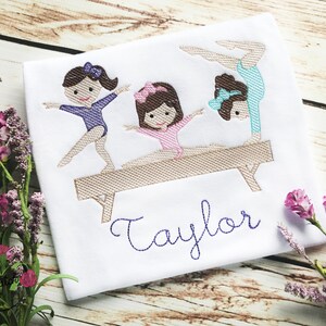 Puede incluir: Dise&ntilde;o bordado de tres ni&ntilde;as haciendo gimnasia en una viga de equilibrio. El dise&ntilde;o incluye el nombre "Taylor" en hilo morado.