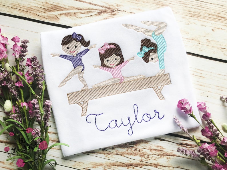 Puede incluir: Dise&ntilde;o bordado de tres ni&ntilde;as haciendo gimnasia en una viga de equilibrio. El dise&ntilde;o incluye el nombre "Taylor" en hilo morado.