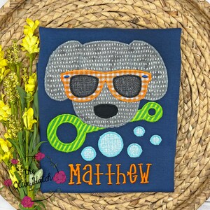 May include: A blue t-shirt with a gray dog wearing orange and white sunglasses and holding a green and yellow bubble wand. There are blue bubbles around the dog and the name "Matthew" is embroidered in orange on the shirt.