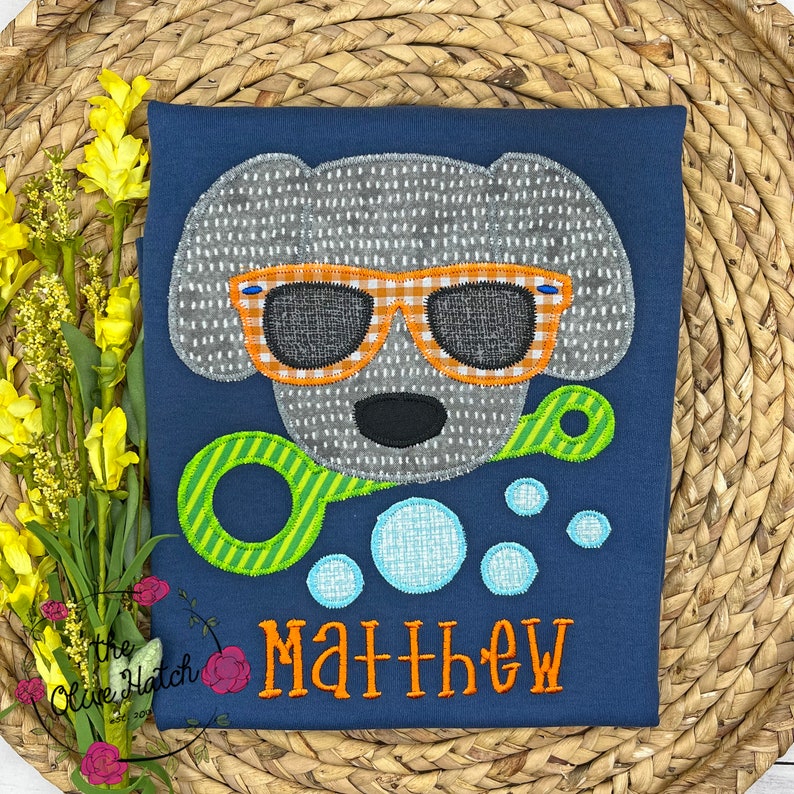May include: A blue t-shirt with a gray dog wearing orange and white sunglasses and holding a green and yellow bubble wand. There are blue bubbles around the dog and the name "Matthew" is embroidered in orange on the shirt.