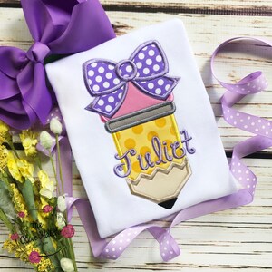 May include: White t-shirt with an embroidered pencil design. The pencil is yellow with a pink eraser and a purple polka dot bow. The name "Juliet" is embroidered in cursive. A large purple bow and ribbon are in the background.