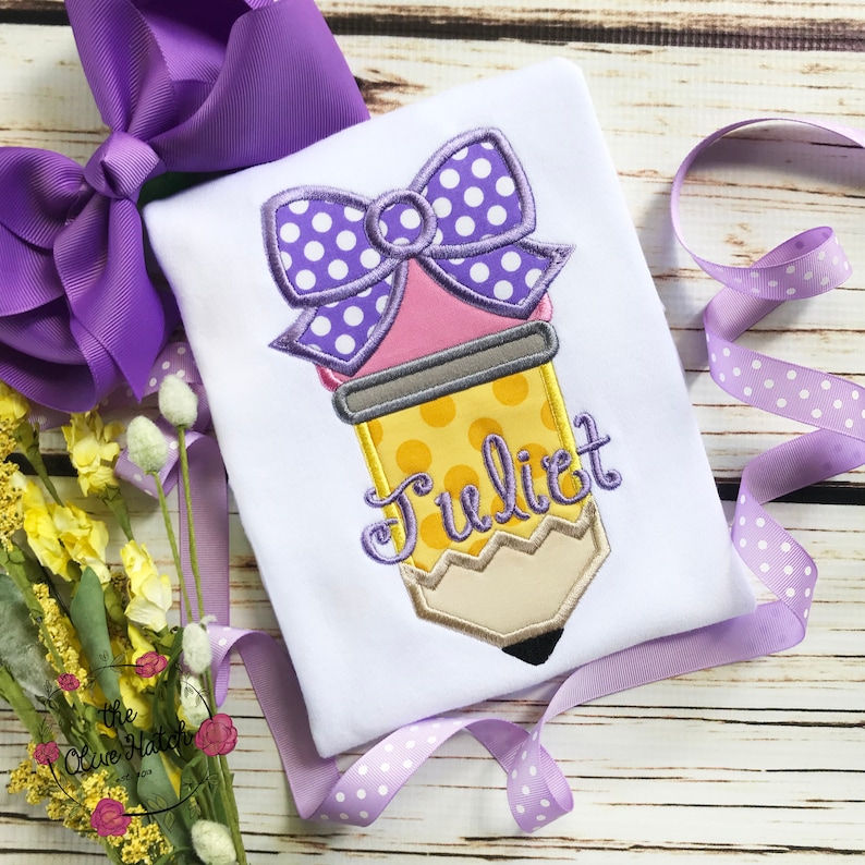 May include: White t-shirt with an embroidered pencil design. The pencil is yellow with a pink eraser and a purple polka dot bow. The name "Juliet" is embroidered in cursive. A large purple bow and ribbon are in the background.