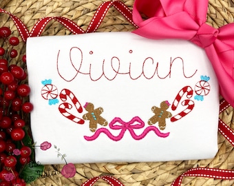 May include: White t-shirt embroidered with the name "Vivian" in red script. The design includes a candy cane, gingerbread man, and bow motif in red, brown, and pink. A large pink bow is placed on the upper right.