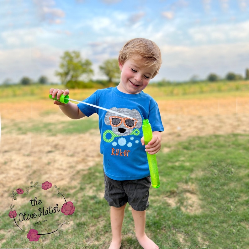 May include: A young boy wearing a blue t-shirt with a cartoon dog wearing sunglasses and the name "Ryker" on it. He is holding a green bubble wand and blowing bubbles in a grassy field.