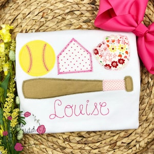 Puede incluir: Camiseta blanca con dise&ntilde;os de b&eacute;isbol bordados en rosa y marr&oacute;n, incluyendo una pelota de b&eacute;isbol, un bate, un guante y un plato de home. El nombre "Louise" est&aacute; bordado en rosa debajo de los dise&ntilde;os.