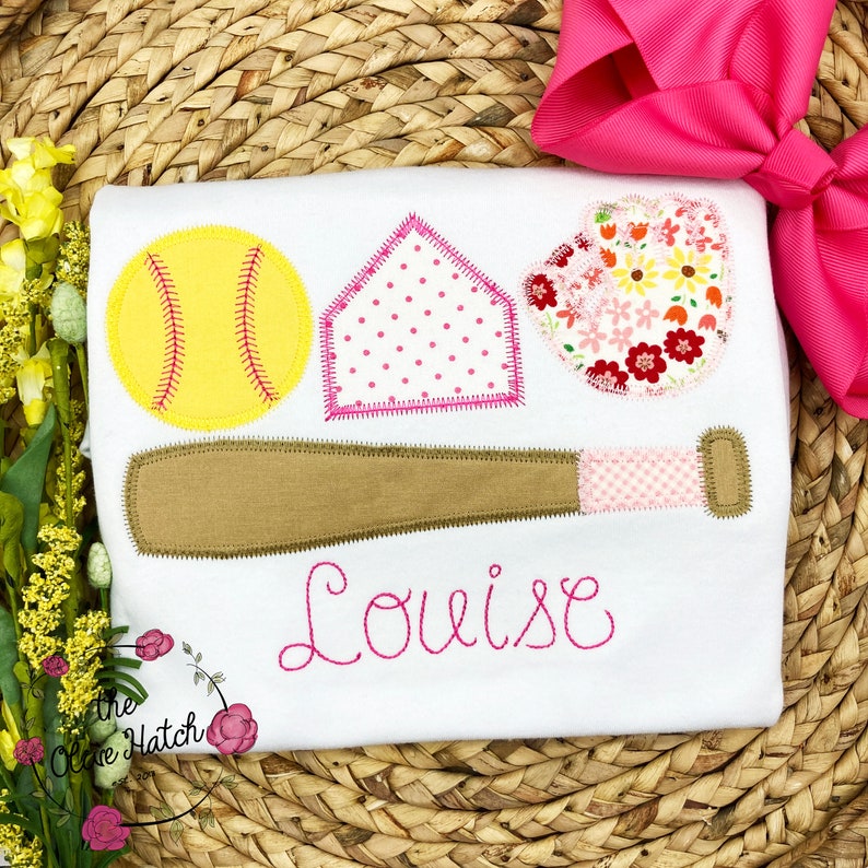 Puede incluir: Camiseta blanca con dise&ntilde;os de b&eacute;isbol bordados en rosa y marr&oacute;n, incluyendo una pelota de b&eacute;isbol, un bate, un guante y un plato de home. El nombre "Louise" est&aacute; bordado en rosa debajo de los dise&ntilde;os.