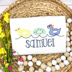 May include: A white onesie with a blue, yellow, and green embroidered design of a duck, donut, and dinosaur in inner tubes. The text "Samuel" is embroidered below the design.