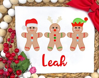 May include: White t-shirt featuring three gingerbread people with festive hats and details. The gingerbread people are decorated with red, green, and blue accents. The name "Leah" is embroidered in red script below the gingerbread figures.
