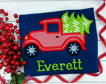 May include: Navy blue shirt with a red gingham truck carrying a green and white striped Christmas tree. The name "Everett" is embroidered in green. A festive design for the holiday season.