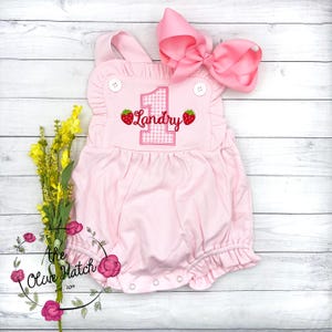 May include: A light pink baby romper with a ruffled neckline and hem. The romper features the number "1" in a pink gingham pattern, the name "Landry", and strawberry appliqués. A large pink bow sits at the top. The Olive Hatch logo is in the bottom left.