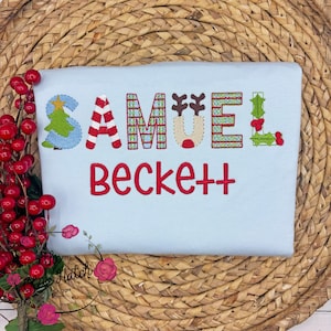 May include: A light blue t-shirt with the name "Samuel Beckett" embroidered in red and festive colors. The letter "S" features a Christmas tree design, and the letters are decorated with candy cane stripes, reindeer antlers, and holly.