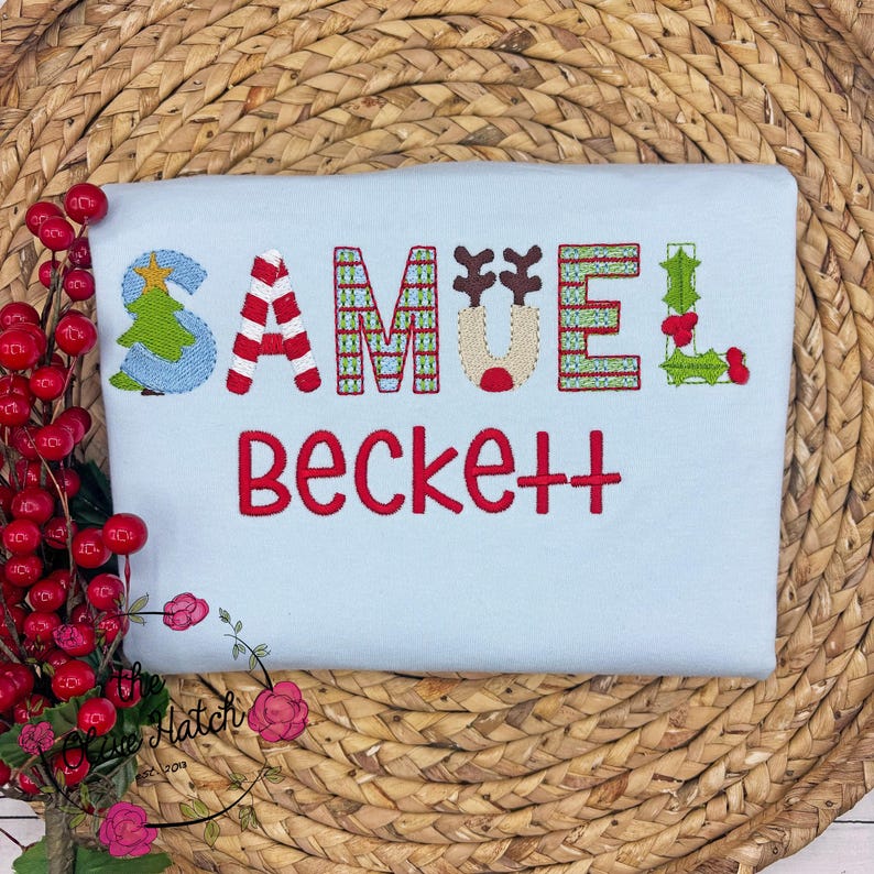 May include: A light blue t-shirt with the name "Samuel Beckett" embroidered in red and festive colors. The letter "S" features a Christmas tree design, and the letters are decorated with candy cane stripes, reindeer antlers, and holly.
