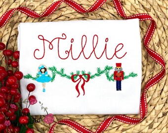 May include: White shirt embroidered with the name "Millie" in red script. Below, a green garland with a blue ballerina and a red nutcracker. A red bow is in the center. A red ribbon with white stitching frames the shirt.