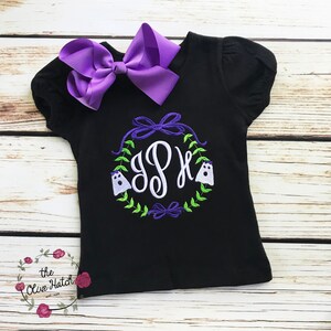 Peut inclure: T-shirt noir &agrave; manches courtes avec un grand n&oelig;ud violet et un motif de couronne. La couronne comprend un monogramme, deux fant&ocirc;mes et des feuilles vertes. Le t-shirt a un col rond et un n&oelig;ud d&eacute;coratif.