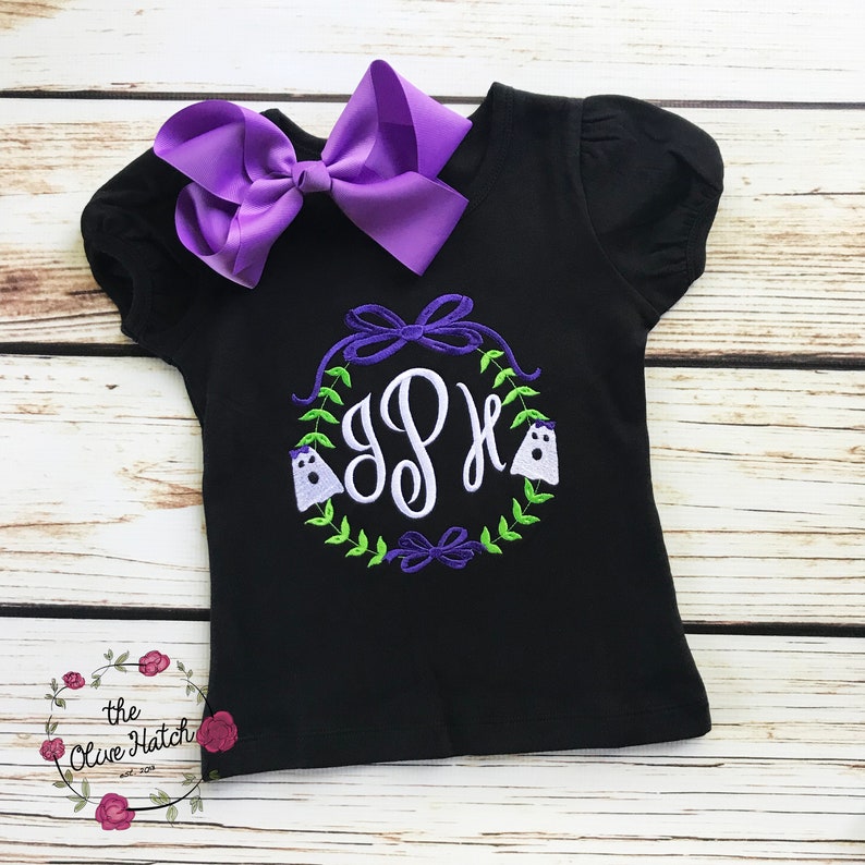Peut inclure: T-shirt noir &agrave; manches courtes avec un grand n&oelig;ud violet et un motif de couronne. La couronne comprend un monogramme, deux fant&ocirc;mes et des feuilles vertes. Le t-shirt a un col rond et un n&oelig;ud d&eacute;coratif.