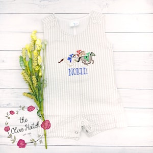 May include: A white and beige striped baby onesie with a horse racing embroidery design and the name "Nolan" embroidered in blue thread.