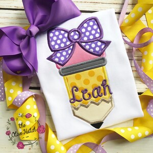 May include: White t-shirt with a pencil design. The pencil is yellow with a pink eraser and a purple bow with white polka dots. The name "Leah" is embroidered in purple. A large purple bow and yellow ribbon with white polka dots are in the background.