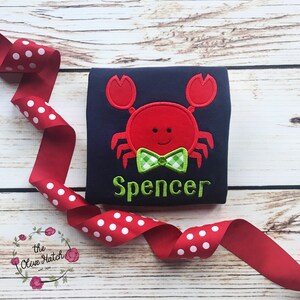 Puede incluir: Camiseta azul marino con un dise&ntilde;o de cangrejo rojo y una pajarita a cuadros verdes y blancos. El nombre "Spencer" est&aacute; bordado en verde. Una cinta roja con lunares blancos en primer plano.