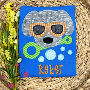 May include: A blue t-shirt with a gray dog wearing orange sunglasses and holding a green and yellow bubble wand. The dog is surrounded by blue and white bubbles. The name "Ryker" is embroidered in orange on the shirt.
