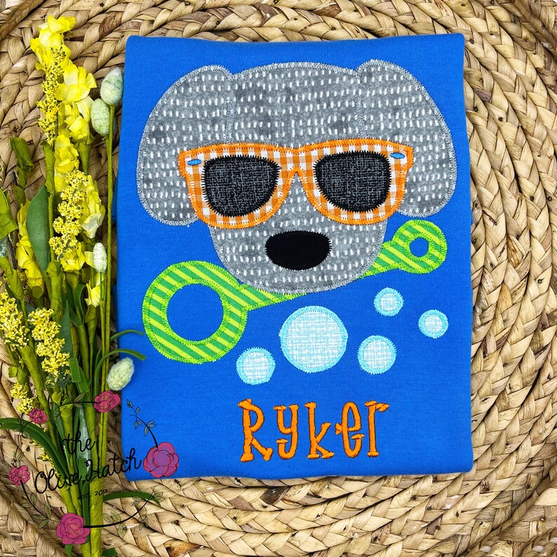 May include: A blue t-shirt with a gray dog wearing orange sunglasses and holding a green and yellow bubble wand. The dog is surrounded by blue and white bubbles. The name "Ryker" is embroidered in orange on the shirt.