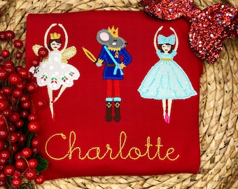 May include: A red long-sleeve shirt featuring embroidered characters from The Nutcracker ballet. The design includes a ballerina, a mouse king, and another ballerina, with the name "Charlotte" embroidered in gold thread. A red glitter bow is in the background.