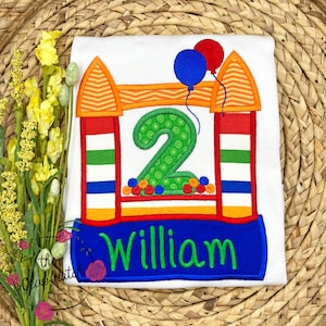 May include: White t-shirt with an embroidered design of a bouncy house with the number 2 and the name William. The bouncy house is decorated with red, orange, green, and blue colors.