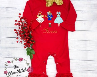 May include: Red baby romper with long sleeves, ruffle cuffs, and a gold bow. The romper features embroidered Nutcracker characters and the name "Olivia". A sprig of red berries and a floral logo are also present.