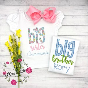 May include: Two white t-shirts with embroidered text. The first shirt says "big sister Annemarie" with floral letters. The second shirt says "big brother Rory" with blue and green letters.