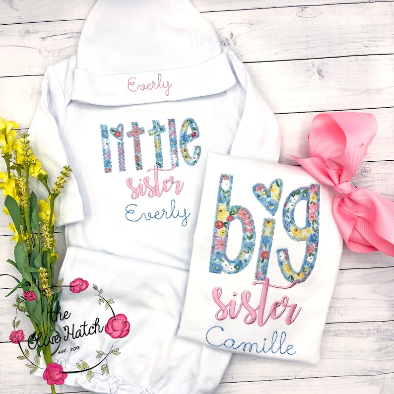 May include: Two white onesies with floral print letters that say "little sister Everly" and "big sister Camille". A pink bow and a white hat with the name "Everly" embroidered on it are also included.