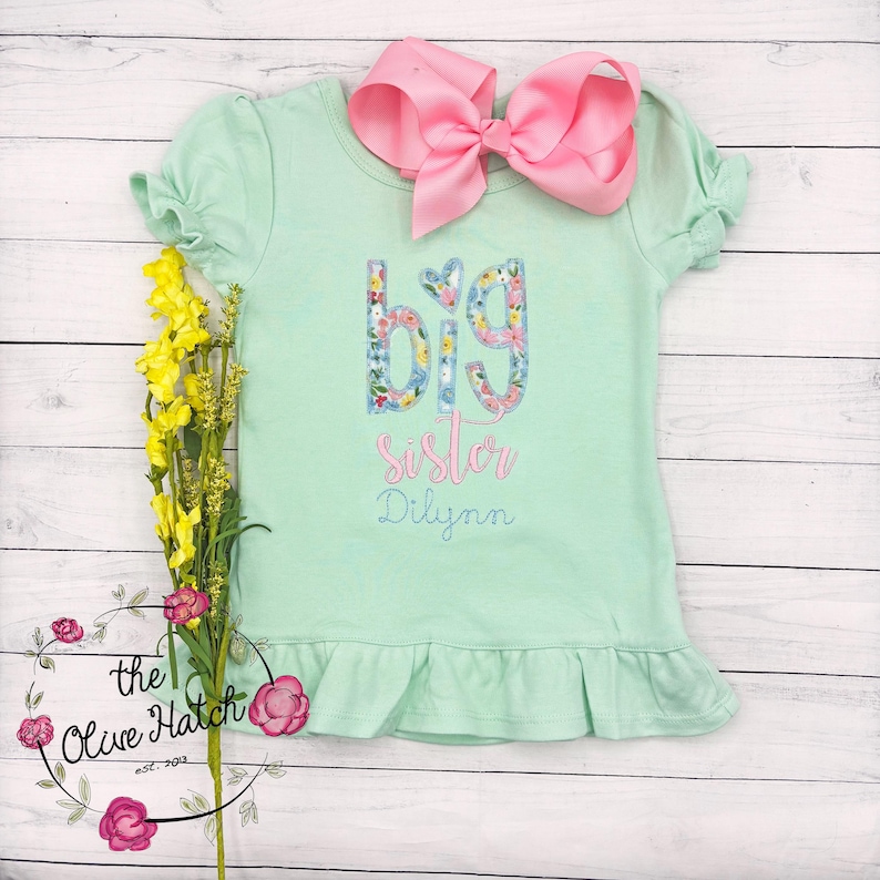 May include: A light green t-shirt with a ruffled hem. The shirt has a floral design that spells out "big sister" with the name "Dilynn" underneath.