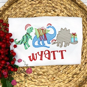 May include: White t-shirt featuring embroidered Christmas dinosaurs. One is green with lights, another blue with a tree, and a third gray with gifts. The name "Wyatt" is embroidered in red below the dinosaurs.