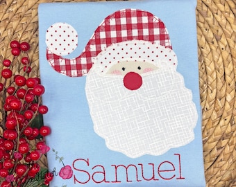 May include: A light blue shirt featuring an embroidered Santa Claus design. The Santa Claus has a red and white checkered hat, a white beard, and a red nose. The name "Samuel" is embroidered in red script below the Santa Claus design.