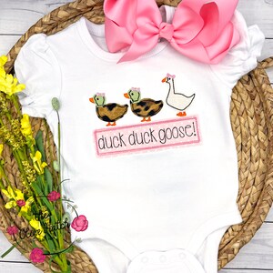Op de afbeelding: Een witte baby romper met een roze strik erboven. De romper heeft een geborduurd ontwerp van twee eenden en een gans met de tekst "duck duck goose!"